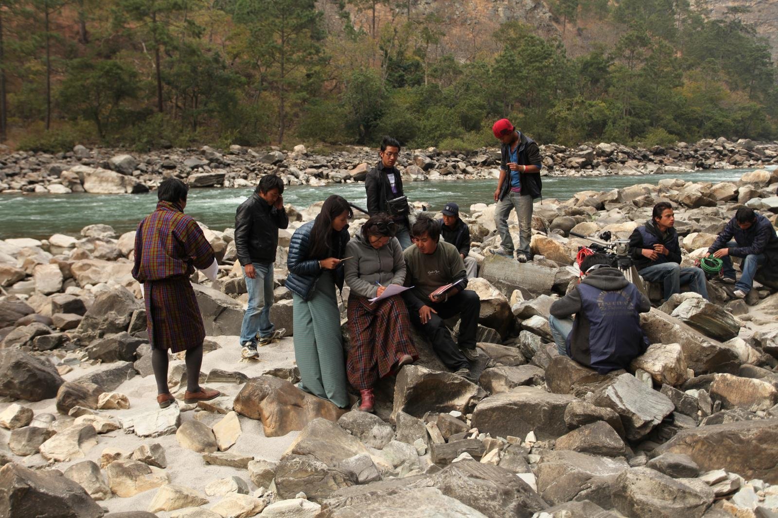 Karma Deki seated on boulders examining script together, while others with camera equipment rest near a turquoise river under a pine-forested hill.